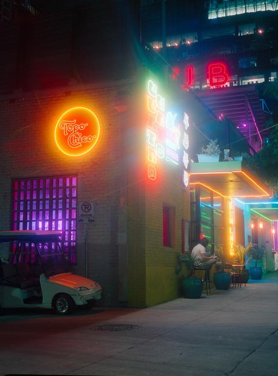 A golf cart is parked next to a building with neon signs and a man sitting at a table on the sidewalk at night.