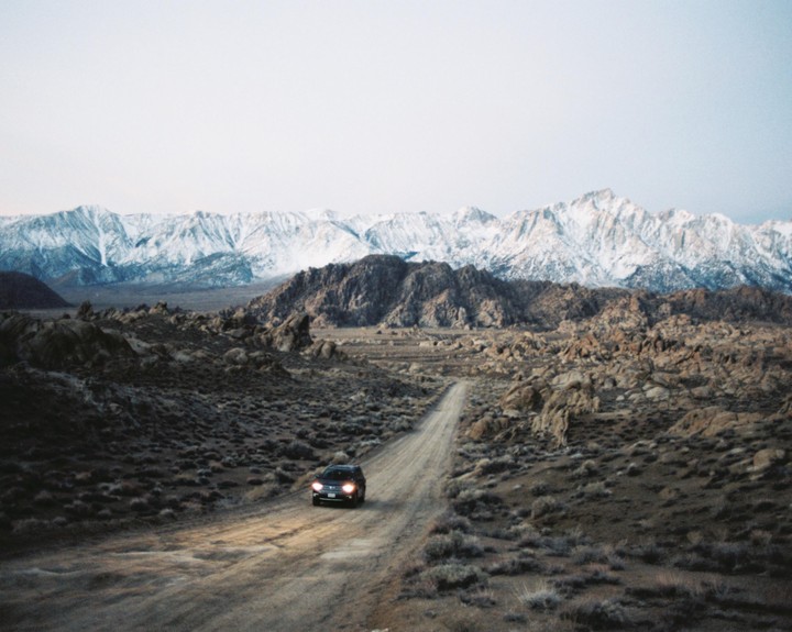 A car drives on a dirt road through a rocky, arid landscape with snow-capped mountains in the distance.