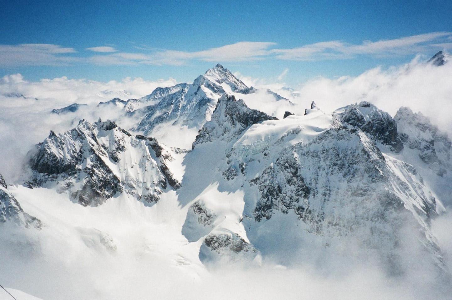 Snow-covered mountains rise above a sea of clouds under a blue sky.