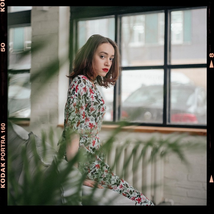 A woman in a floral dress sits by a window with green foliage in the foreground.