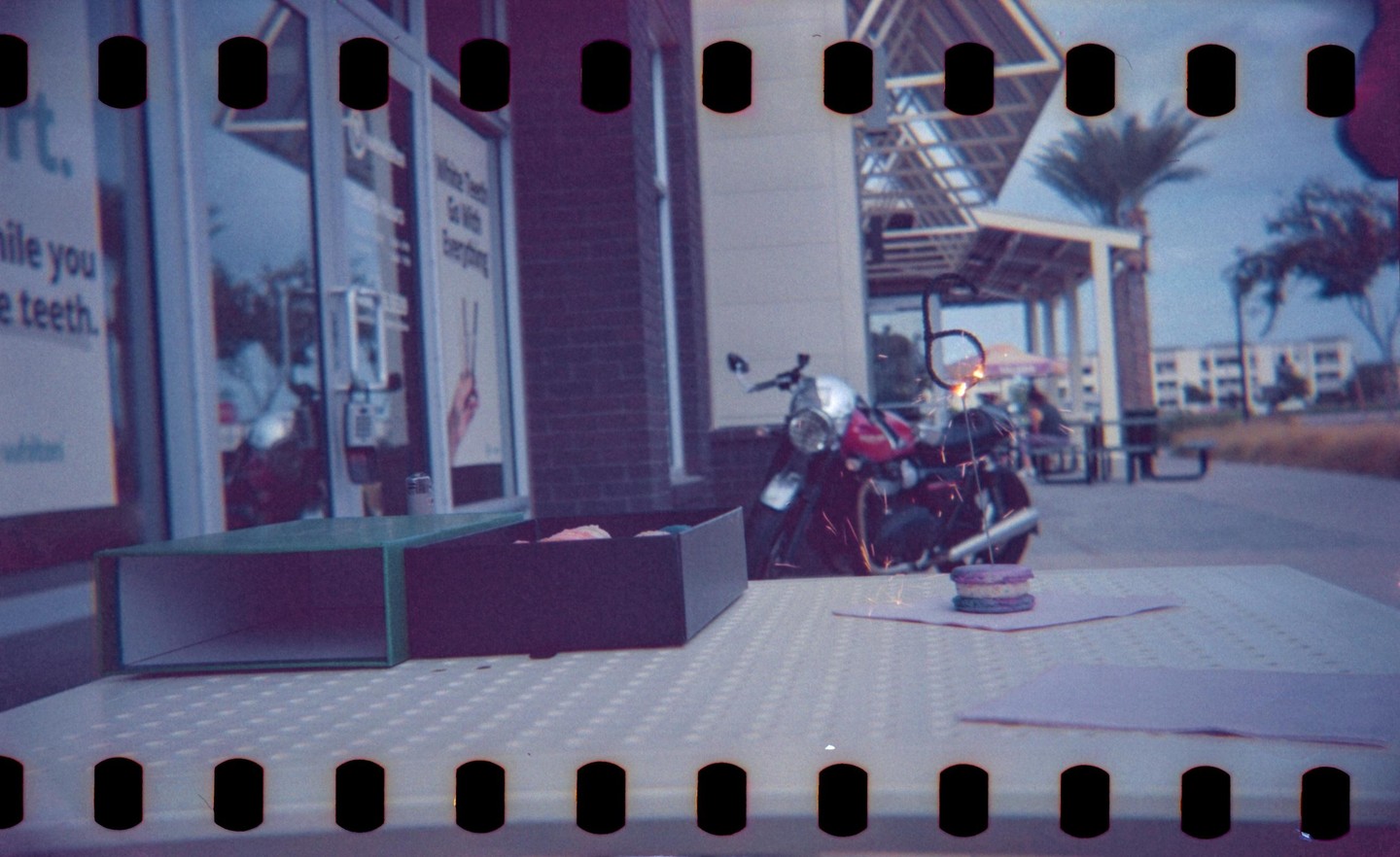 A motorcycle is parked outside a building with a table in the foreground holding macarons and a sparkler.