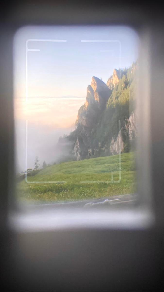 A mountain landscape with fog and green grass is visible through a camera viewfinder.