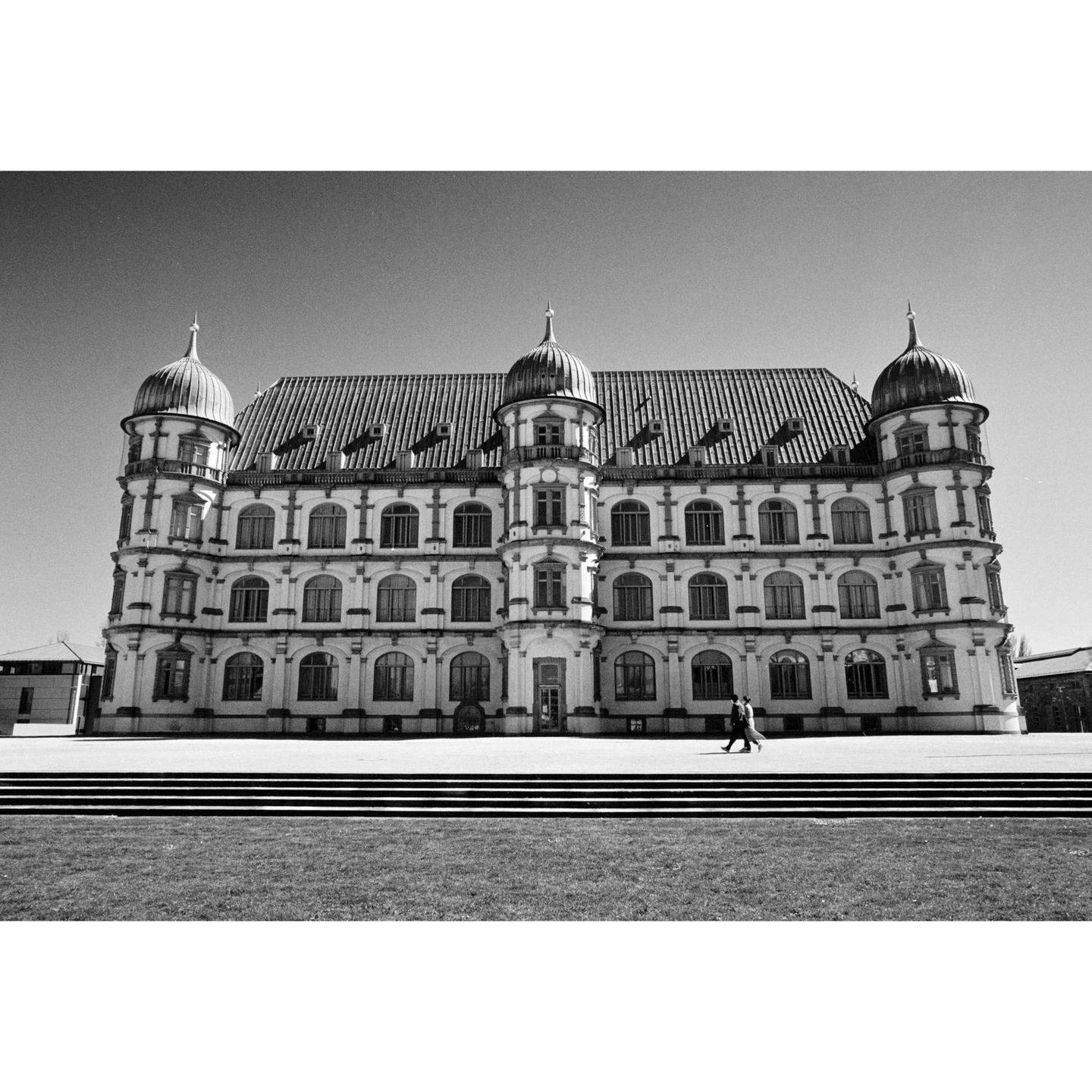A large ornate building with multiple domes and arched windows stands behind a wide set of steps and a grassy area.