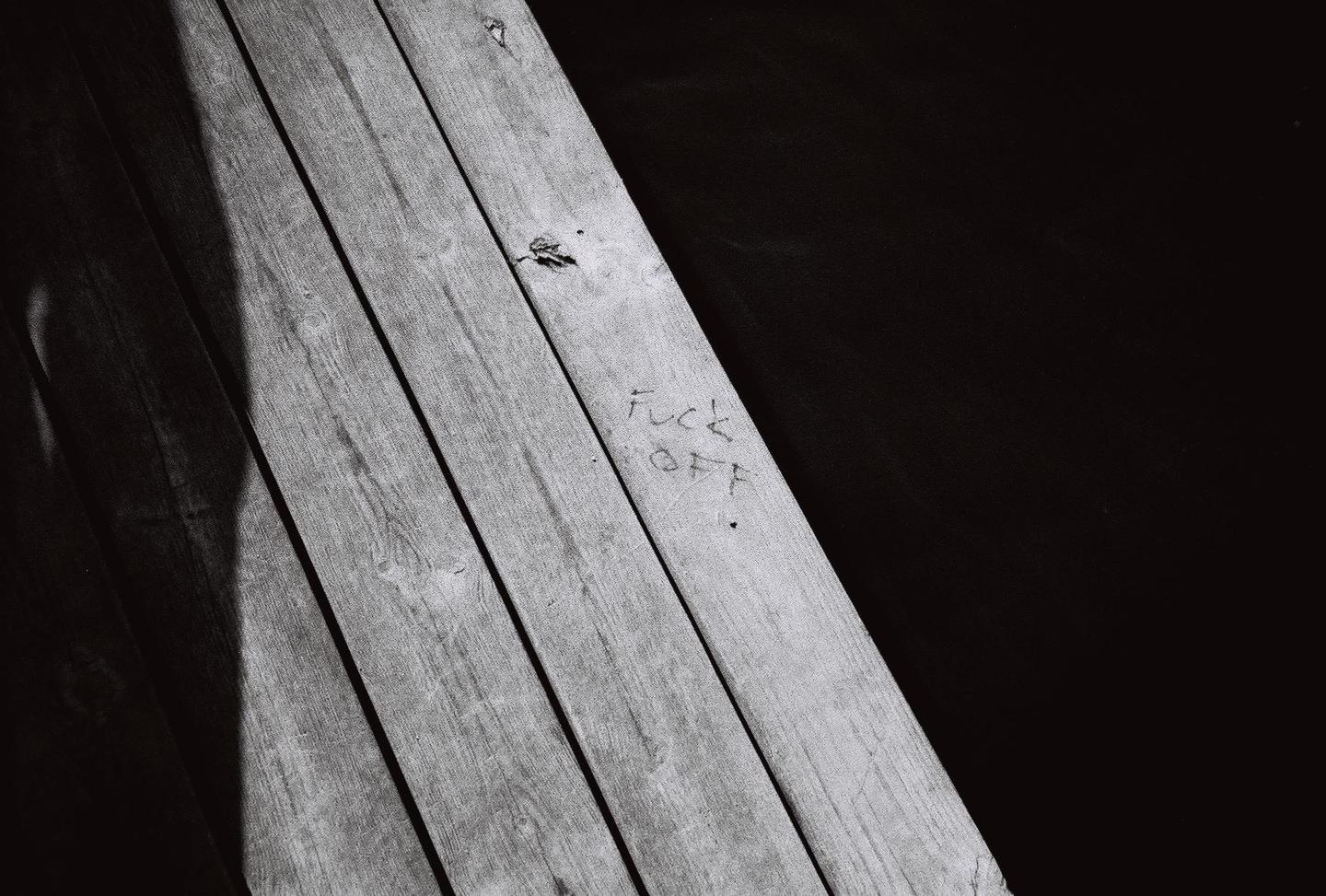 The words "Fuck Off" are carved into a wooden plank next to a dark shadow.