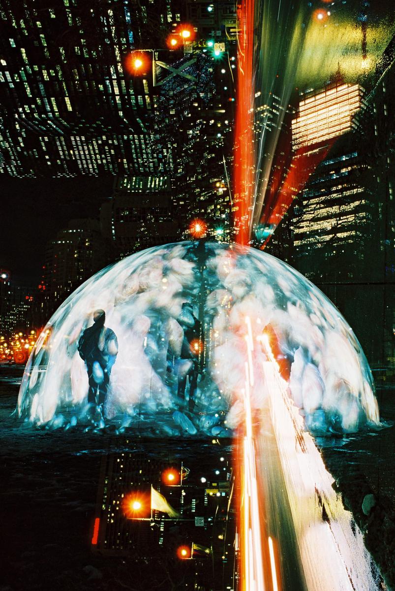 A man stands inside a glowing dome at night with city lights and light trails in the background.