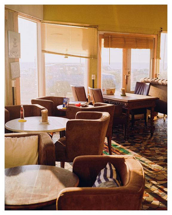 A cafe with several tables and chairs is illuminated by bright sunlight streaming through large windows.
