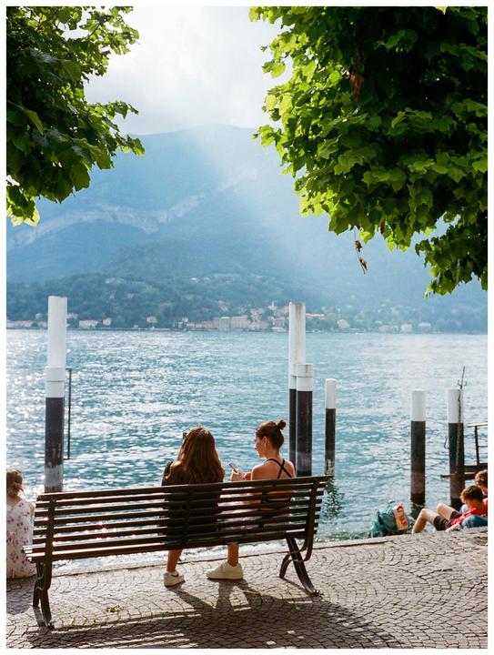 Two women sit on a bench by a lake with mountains in the background.