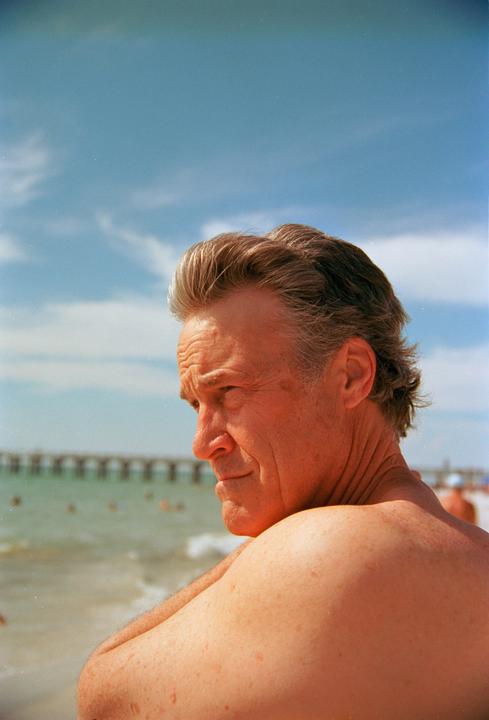 A man with gray hair looks out at the ocean from a beach with a pier in the background.