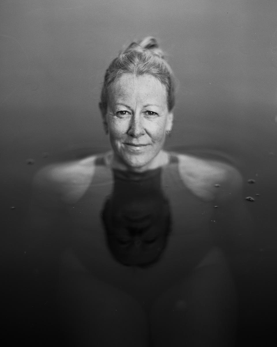 A woman with her hair in a bun is in water, with her reflection visible below her.