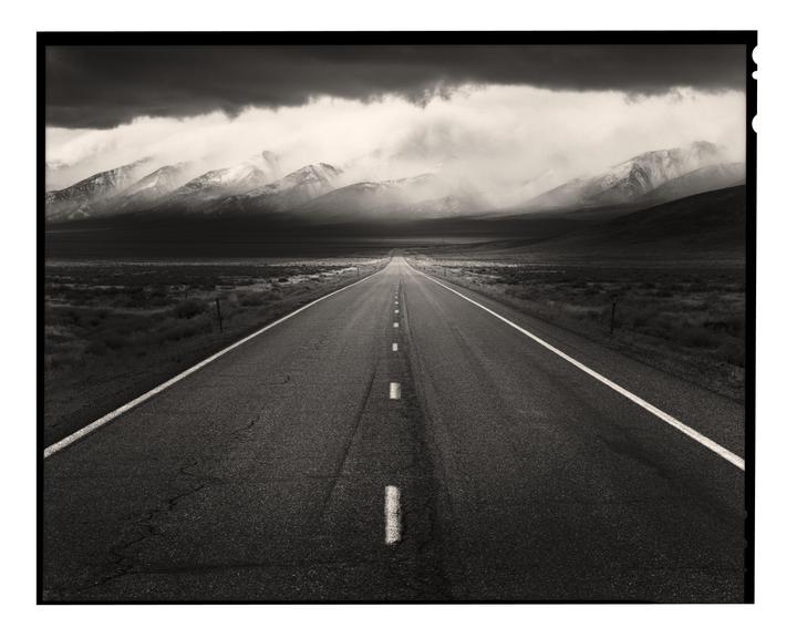 A long, straight road leads towards snow-capped mountains under a dramatic, cloudy sky.