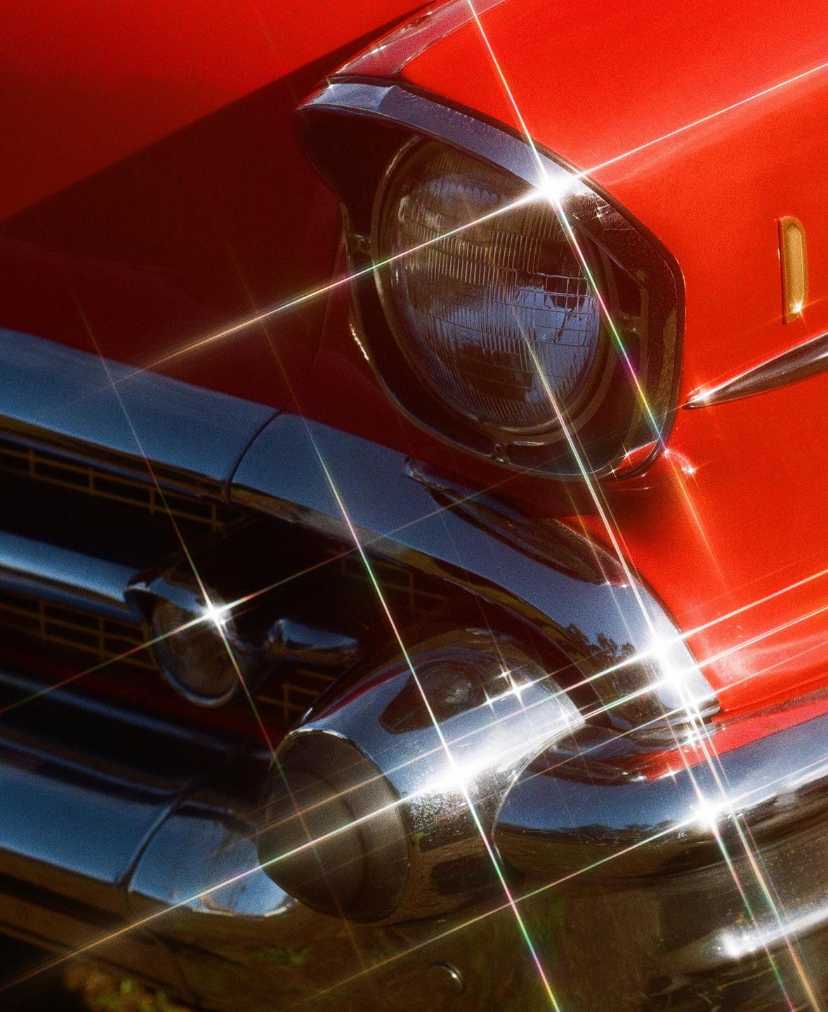 A red car with a shiny headlight and chrome bumper is shown in a close-up.