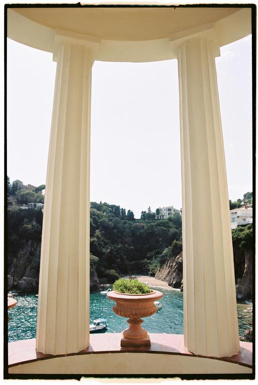 A terracotta planter with green foliage sits on a balcony overlooking a cove with boats and a sandy beach, framed by two large columns.