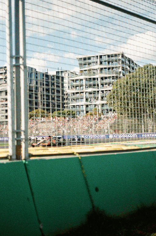 Race cars speed around a track with spectators and buildings in the background.