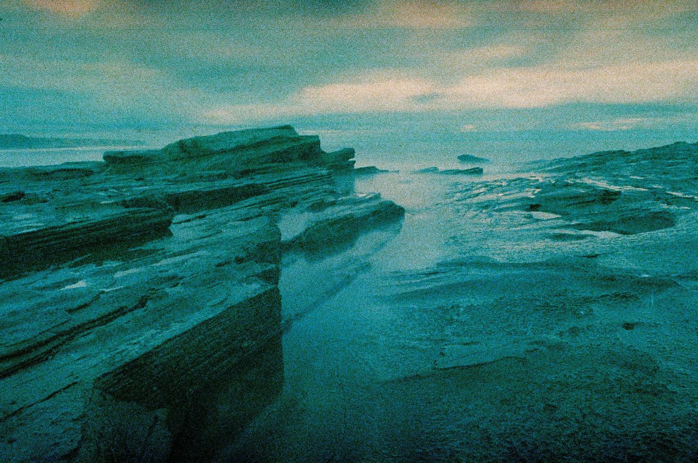 Jagged rock formations line a misty, teal-colored coastline under a cloudy sky.
