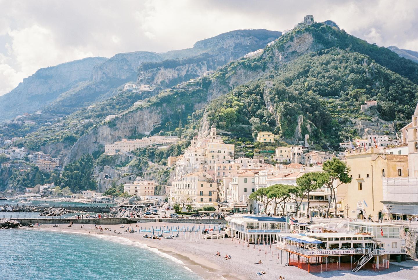 A coastal town with buildings nestled into a mountainside overlooks a beach with people and umbrellas.