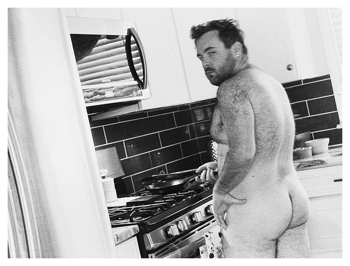 A man stands naked in a kitchen, cooking on a stove.