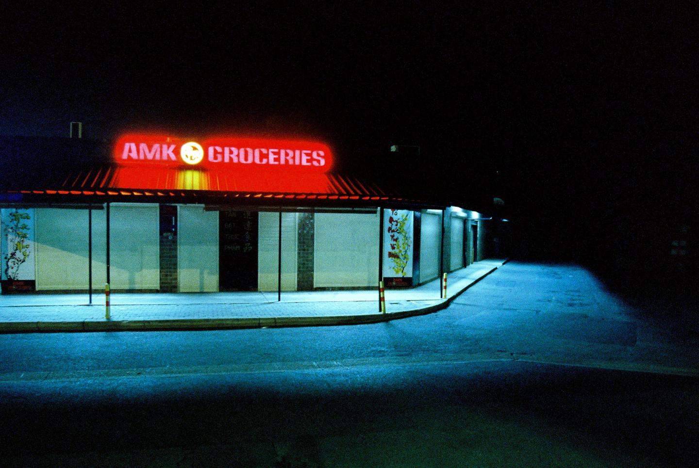 A grocery store with a red neon sign is illuminated at night, casting a blue glow on the street and sidewalk.