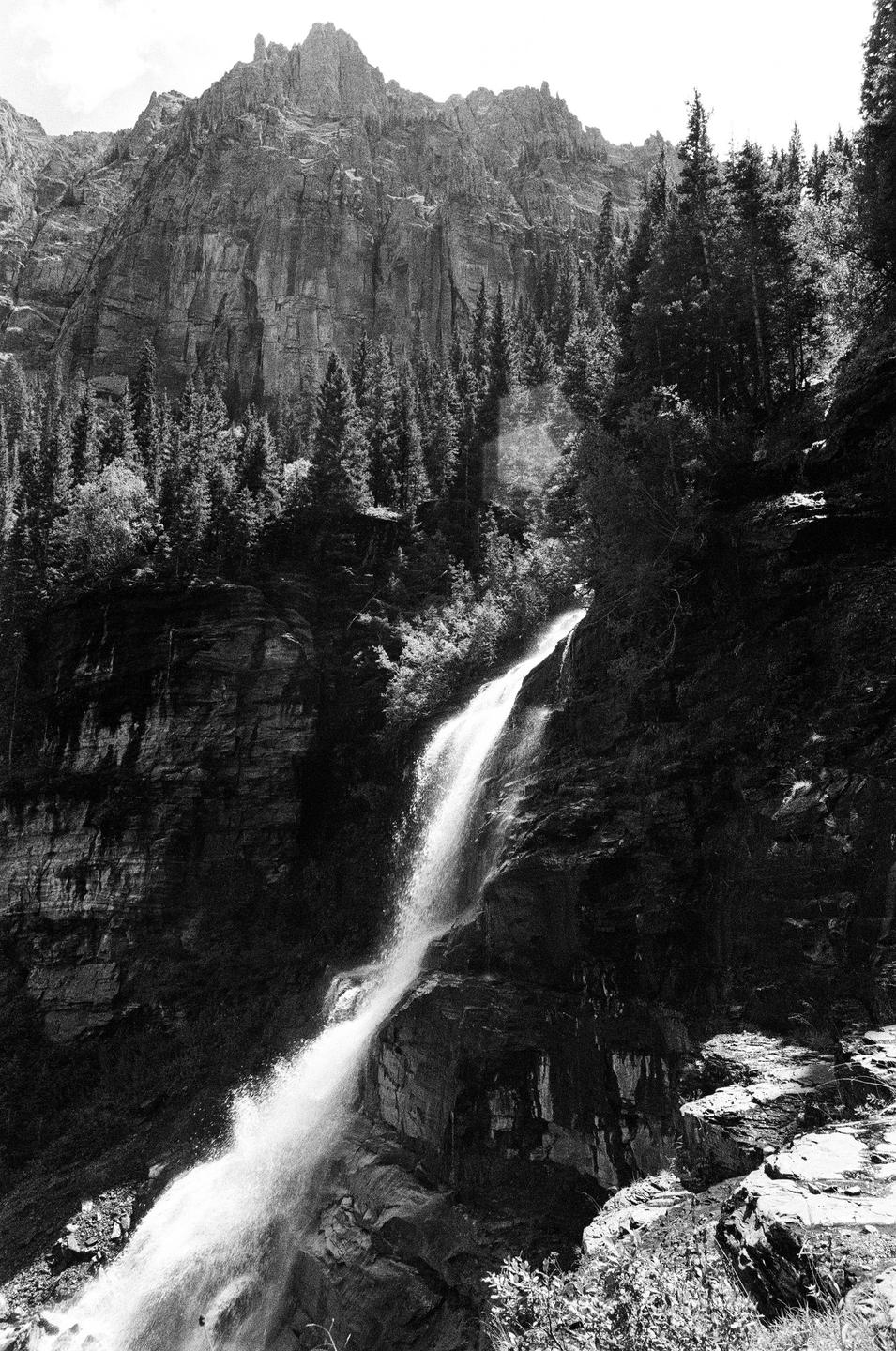 A waterfall cascades down a rocky cliff face surrounded by pine trees and mountains.