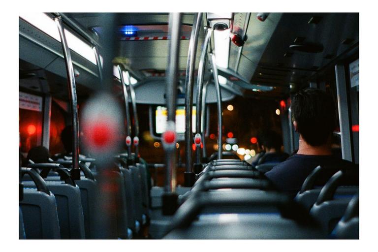 The interior of a bus is visible at night, with a man sitting in the foreground and city lights blurred outside the windows.