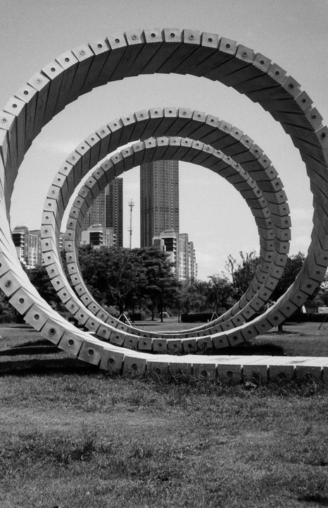 A large spiral sculpture made of concrete blocks stands in a grassy park with tall buildings in the background.
