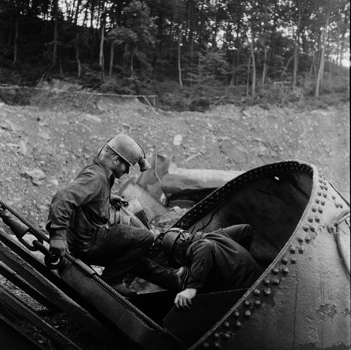 Two miners wearing hard hats and headlamps are working with heavy machinery in a wooded, rocky area.