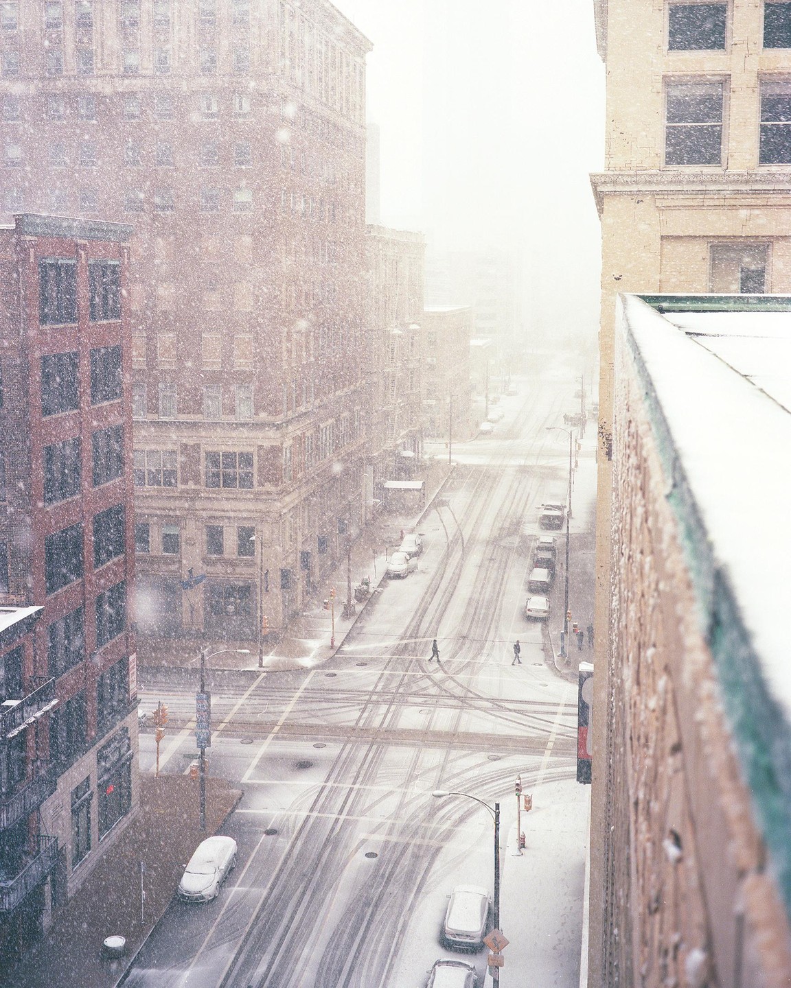 A snow-covered city street with buildings and cars is seen from above during a snowfall.