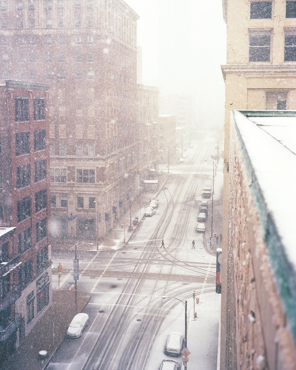 A snow-covered city street with buildings and cars is seen from above during a snowfall.