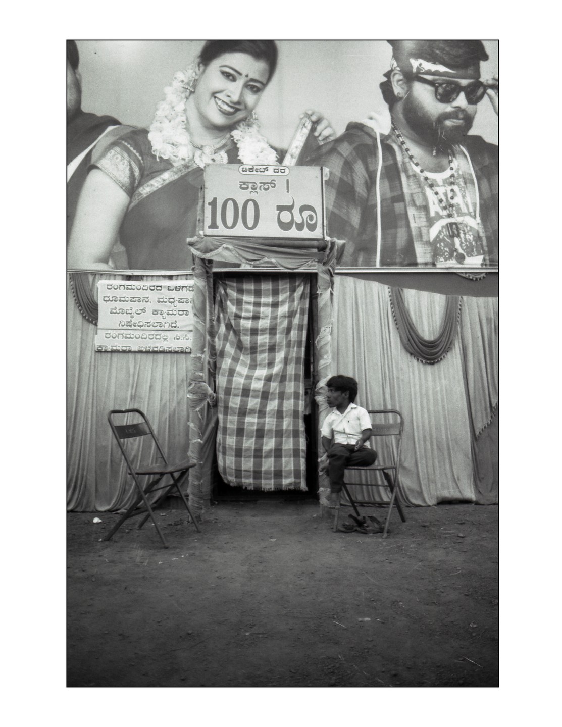 A boy sits on a folding chair in front of a curtained entrance with a large banner of a man and woman above it.