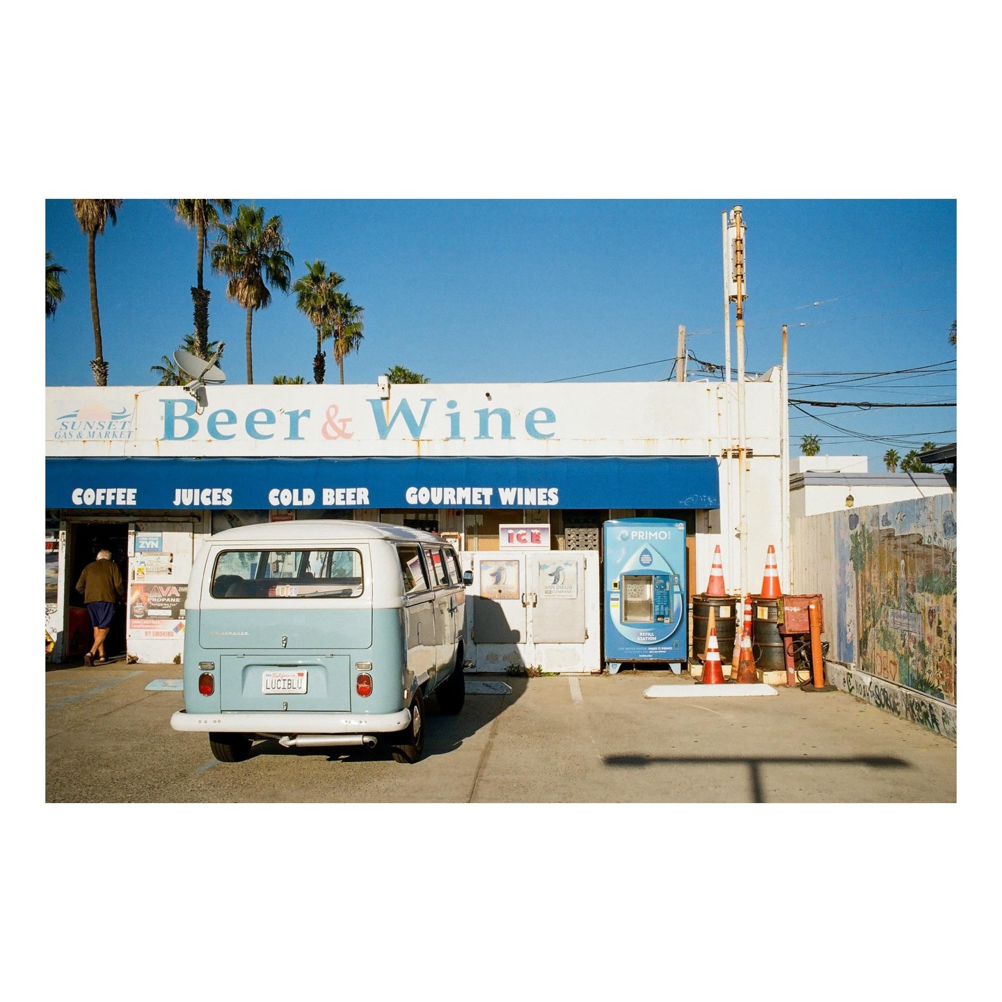 A light blue and white van is parked in front of a store with a blue awning that reads "Beer & Wine" under a clear blue sky.
