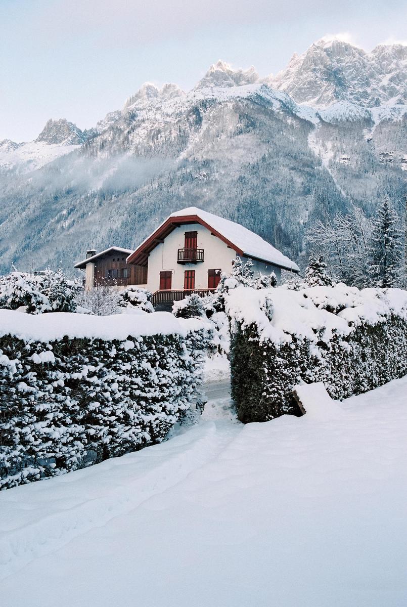 A snow-covered house with a red roof is nestled among snow-laden hedges and trees, with a towering mountain range in the background.