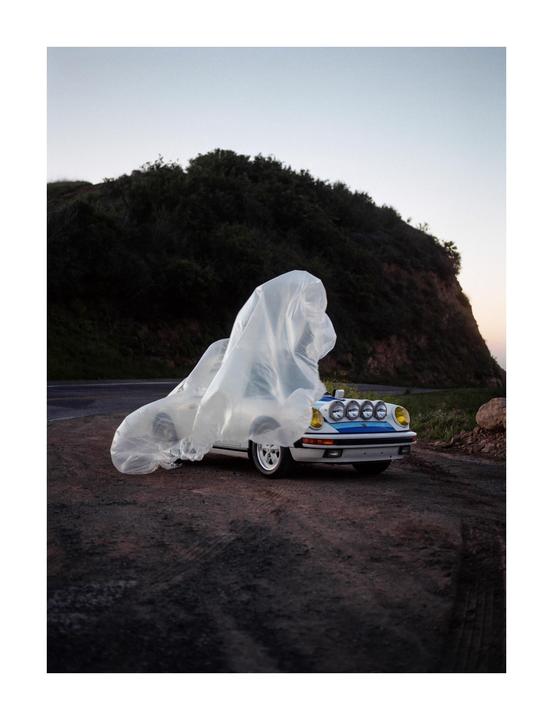 A white car with blue stripes and yellow headlights is parked on a dirt road next to a hill, partially covered by a translucent plastic sheet.