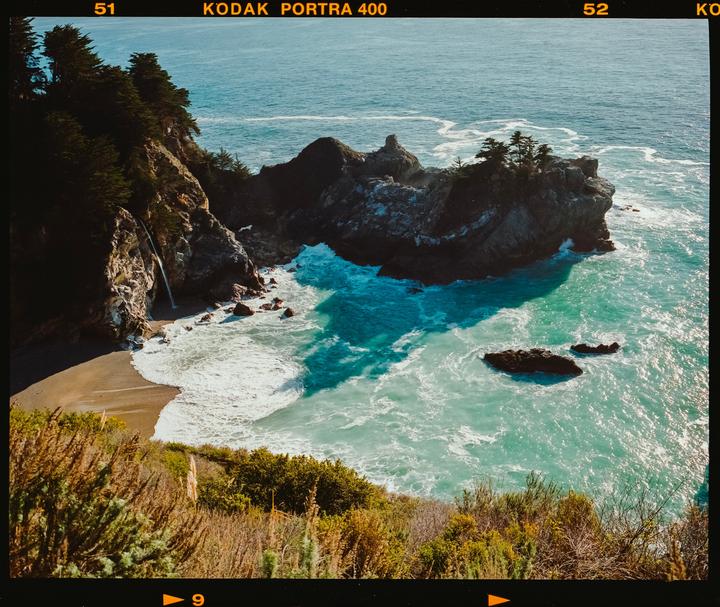 A waterfall cascades onto a sandy beach next to a rocky outcropping in the ocean.