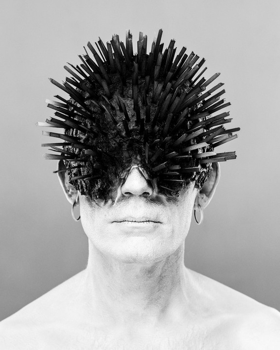 A person with white face paint and a spiky headpiece covers their eyes.