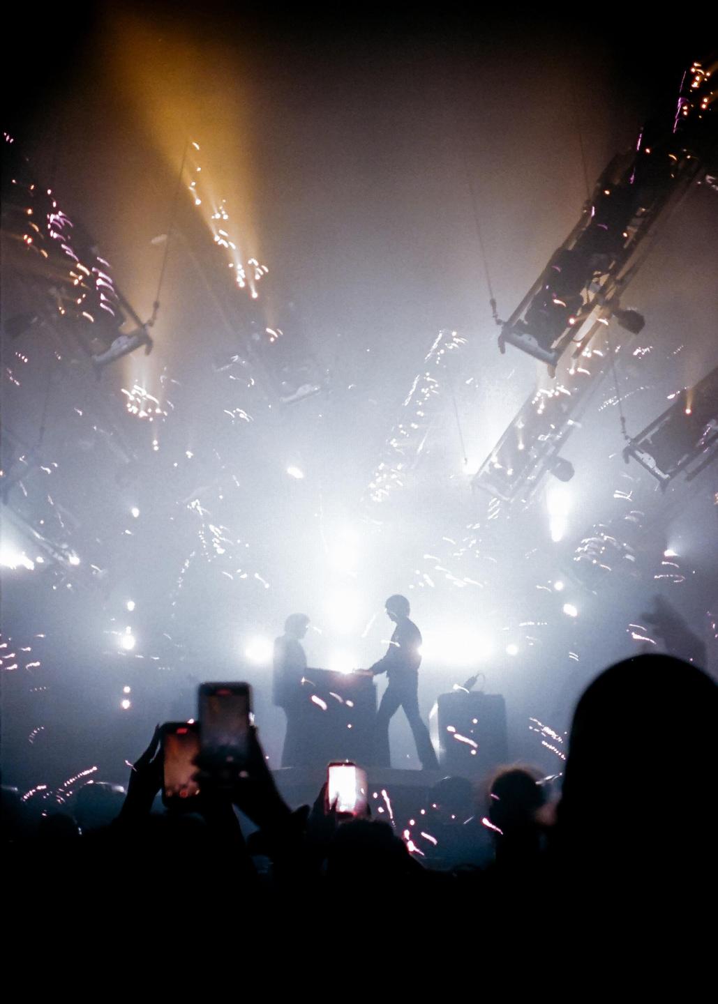 Two men are performing on a stage with bright lights and pyrotechnics while an audience records them with their phones.