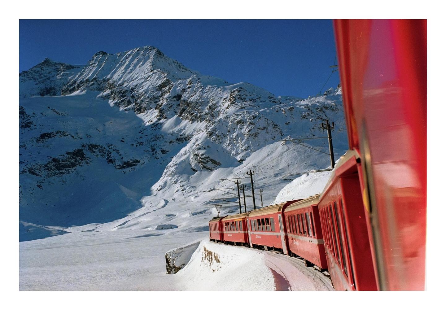 A red train travels along a snowy mountain landscape under a clear blue sky.