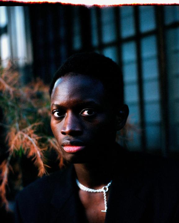 A man with a necklace looks directly at the viewer, with foliage and a window in the background.