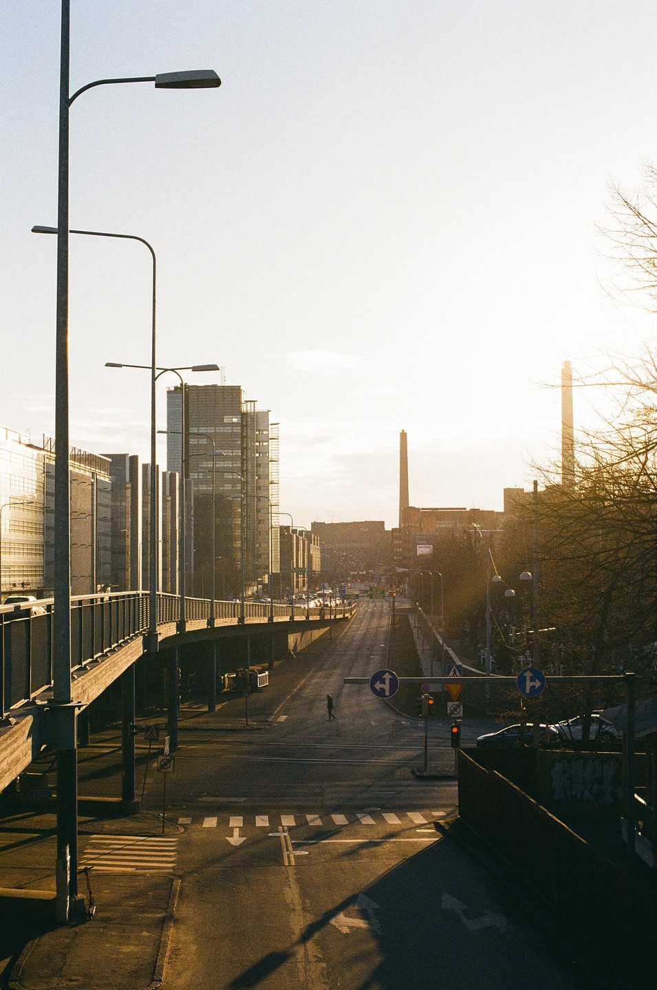 A person walks across a street with an overpass and buildings under a bright sky.
