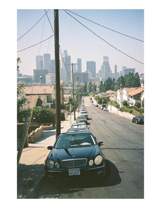 A black car is parked on a residential street with a city skyline in the distance.