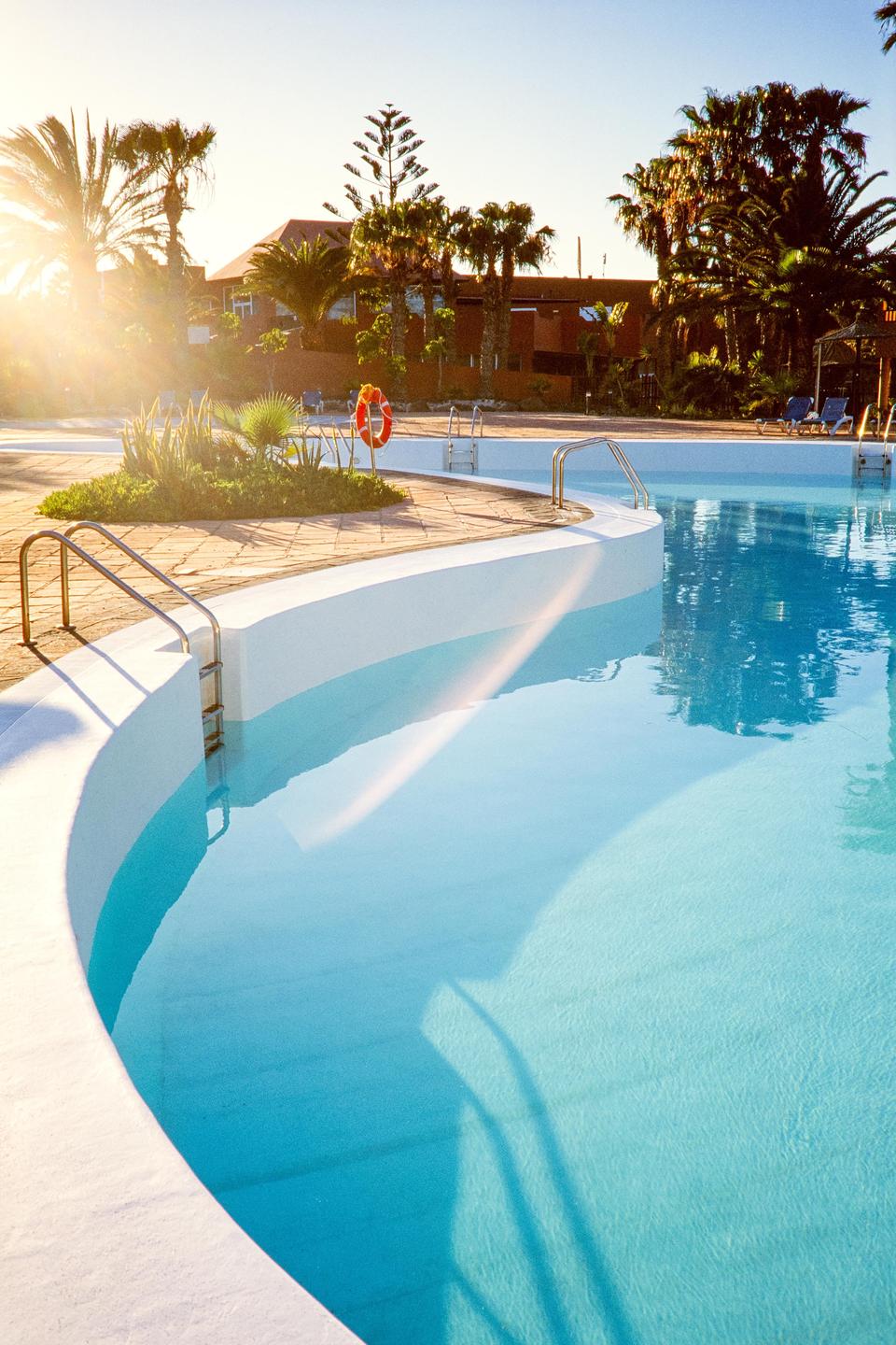 A swimming pool with clear blue water is surrounded by palm trees and buildings under a bright sky.