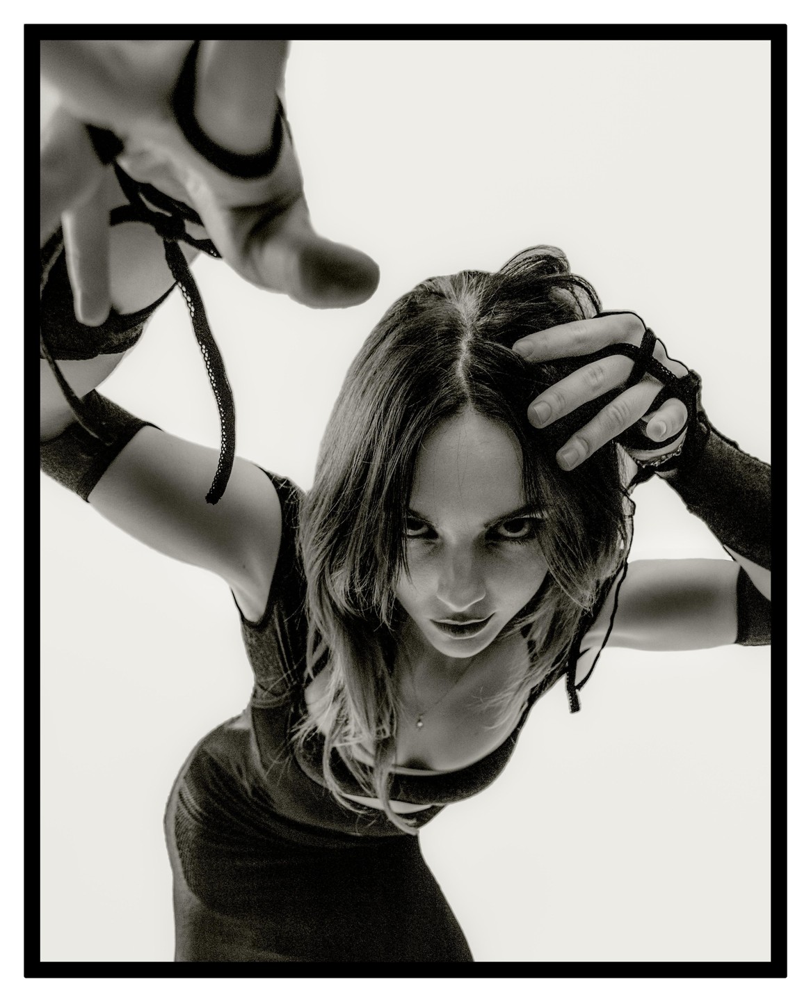 A woman with dark hair and a dark dress looks intensely at the camera, with one hand reaching out towards the viewer and the other in her hair.