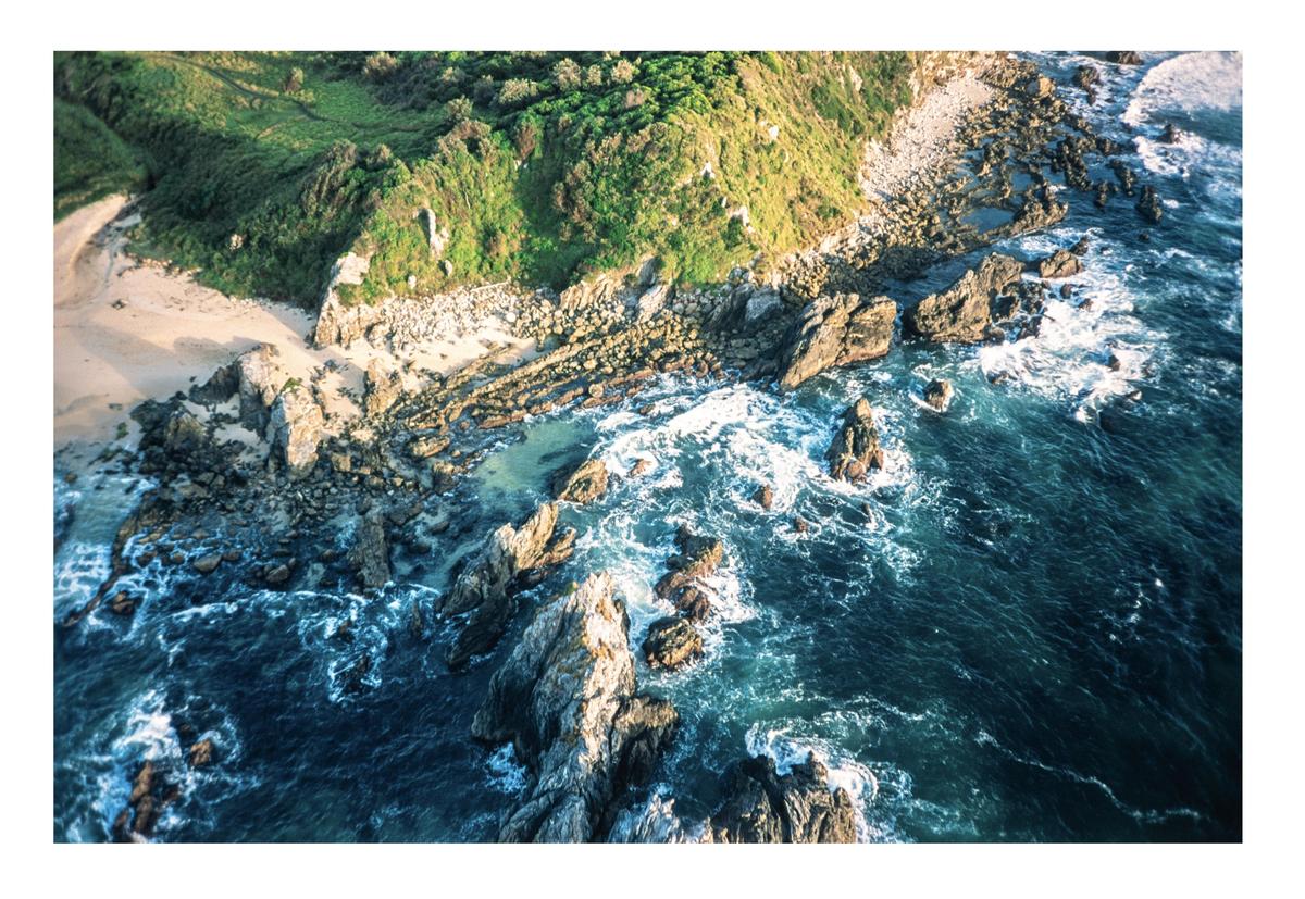 An aerial view shows a rocky coastline with green hills meeting the ocean.