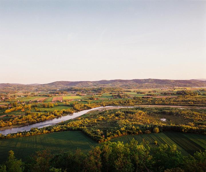 A river winds through a green valley with fields and houses under a clear sky.