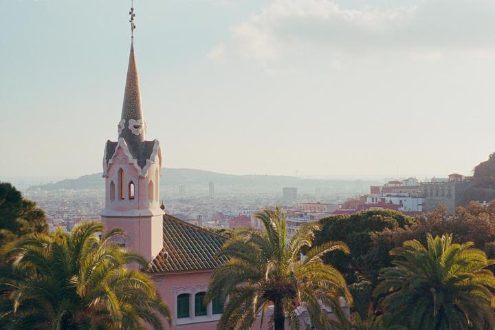 A pink turret with a pointed roof stands above palm trees and a city.