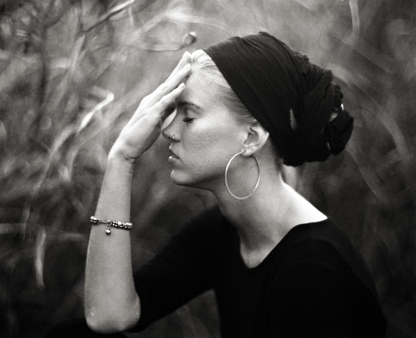 A woman with a headscarf and large hoop earring holds her hand to her forehead with her eyes closed.