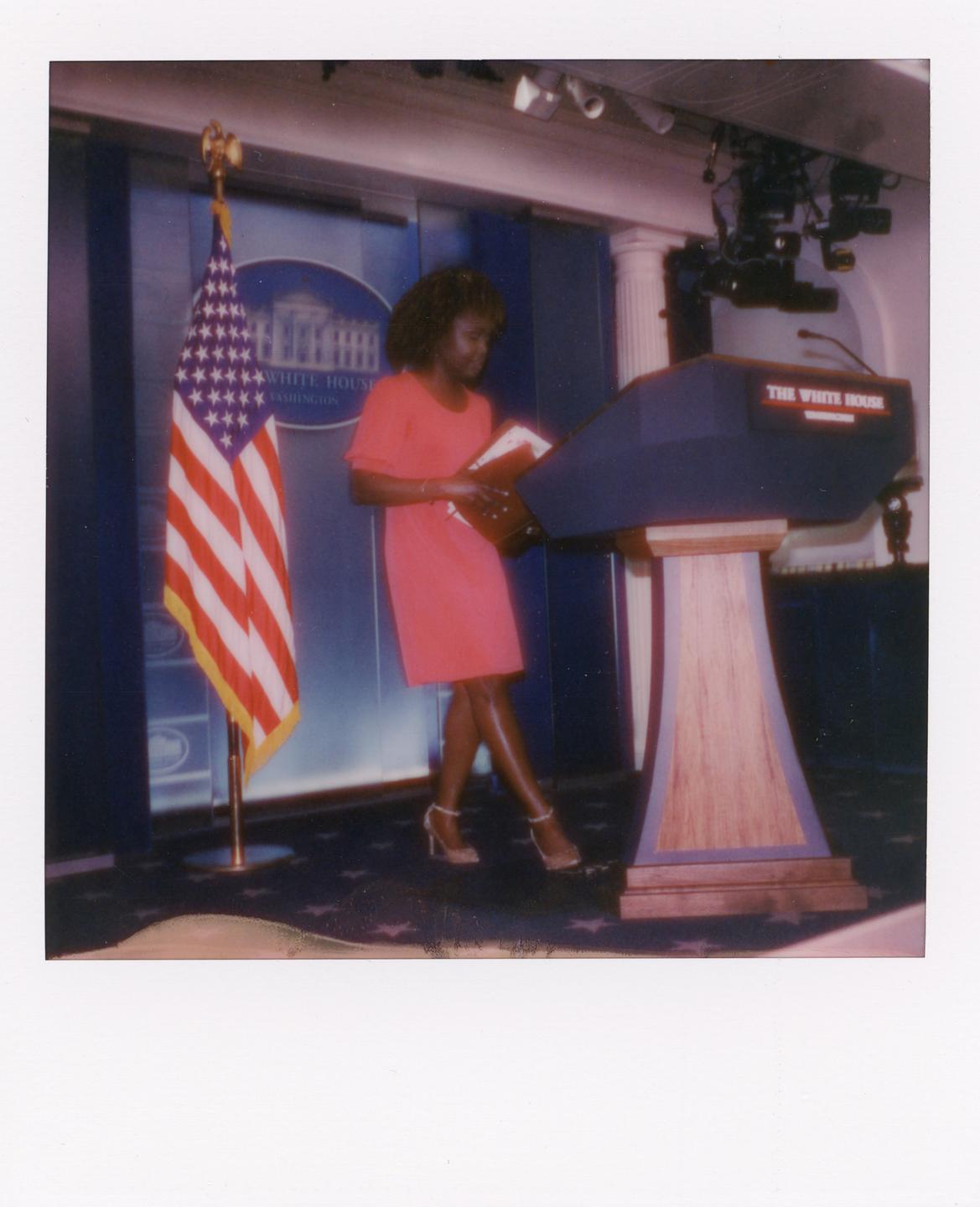 A woman in a red dress stands next to a podium with an American flag in the background.