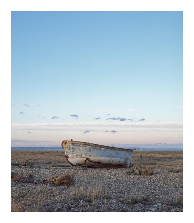 An old, weathered boat rests on a rocky beach under a vast sky with scattered clouds.