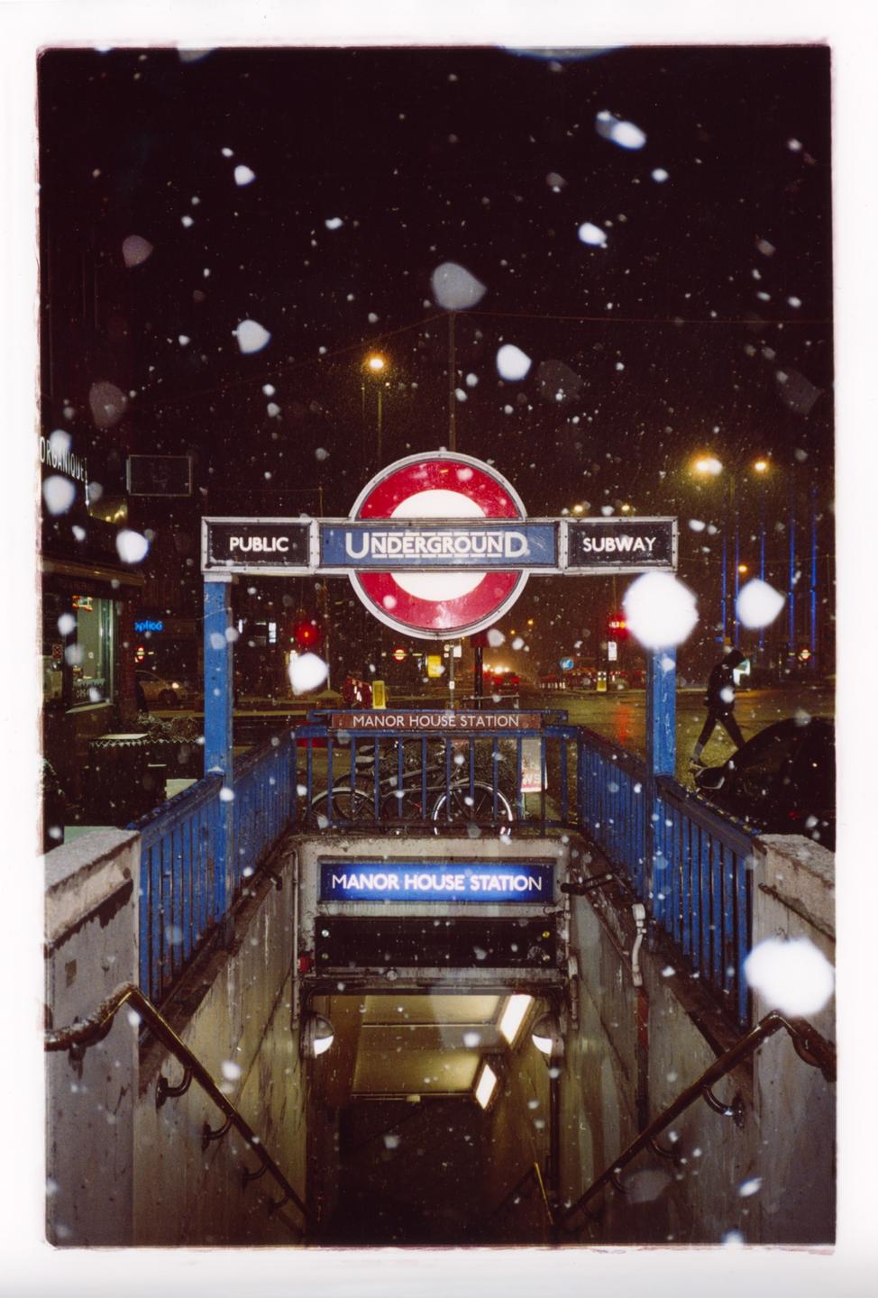 Snow falls around the entrance to Manor House Station in London at night.