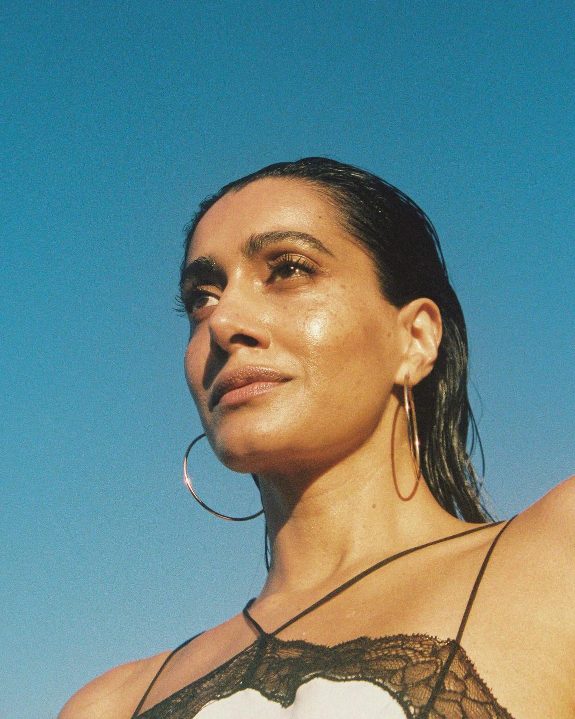 A woman with wet hair and hoop earrings looks up against a clear blue sky.