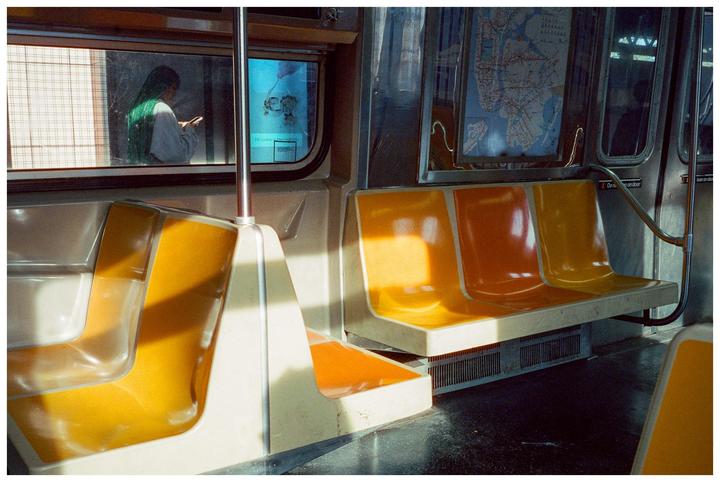A woman with green hair looks at her phone while sitting in a subway car with orange and yellow seats.