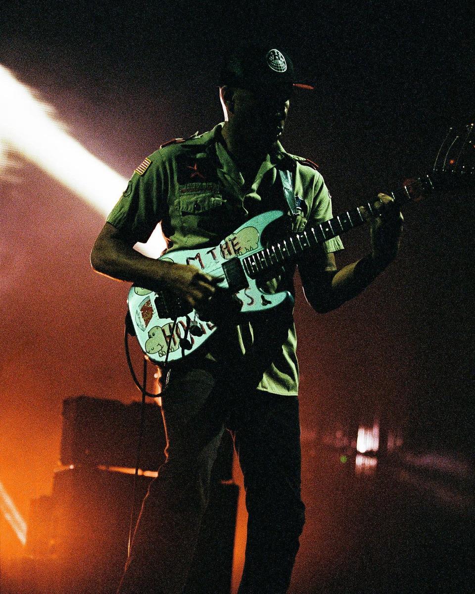 A man in a military-style shirt and cap plays an electric guitar on a stage with orange and white lighting.